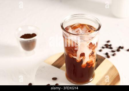 Kaffeeskultur und Morgenroutine, Eiskaffee mit Milch, die in einem Glas auf einer Holzplatte wirbelt, mit gerösteten Kaffeebohnen auf einer weißen strukturierten Oberfläche Stockfoto