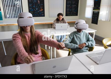 Zwei weibliche Teenager, ein männlicher Teenager, der VR-Headsets in gelben Stühlen verwendet, Laptops im Klassenzimmer. Virtual, Reality, Brillen, Kopfhörer, Geräte, lernen Stockfoto