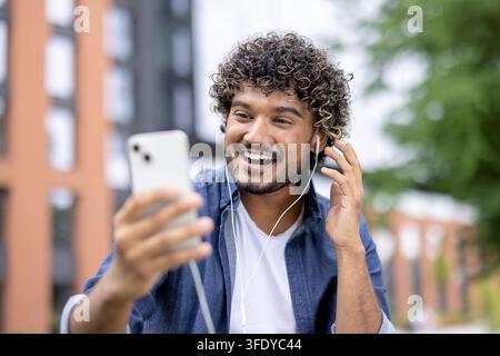 Junger Mann mit lockigem Haar und Kopfhörern lächelt, hat ein Videoanruf auf seinem Smartphone, während er im Freien steht und sich aus der Ferne mit Freunden oder Familie verbindet Stockfoto