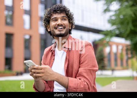 Zufriedener junger Mann mit lockigen Haaren, der im Freien lächelt, ein Smartphone hält und nach oben schaut, und einen Moment der Freizeit in einem modernen Stadtpark mit Gebäuden und Grün genießt Stockfoto