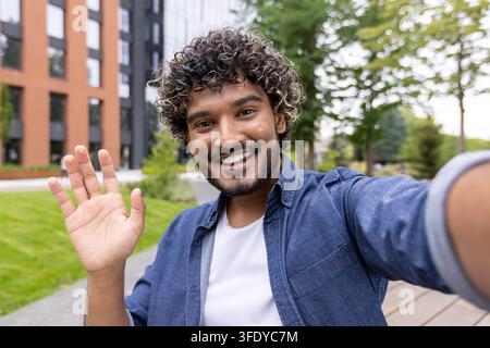 Junger indischer Mann mit lockigem Haar lächelt und winkt mit der Hand, während er im Freien in einem Park mit städtischen Gebäuden im Hintergrund ein Selfie macht, und übermittelt eine freundliche Begrüßung und moderne Kommunikation Stockfoto