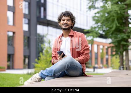 Junger Mann mit lockigen Haaren lächelt und schaut in die Kamera, hält ein Smartphone und sitzt im Freien auf einem Universitätscampus, was moderne Bildung und Technologie repräsentiert Stockfoto