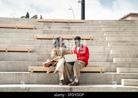 Schwarze Studentenfreunde, die gemeinsam mit einem Laptop auf zeitgenössischen Außentreppen über Kursarbeiten diskutieren Stockfoto