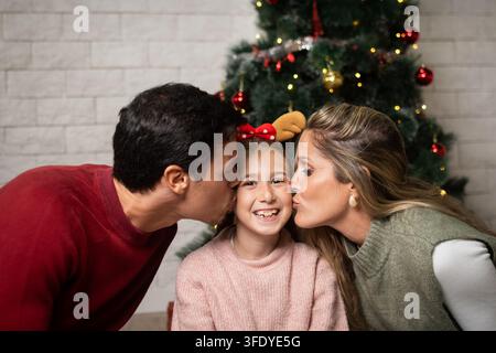 Liebevolle Mutter und Vater küssen ihre lächelnde Tochter auf die Wangen, während sie zu Hause Weihnachten feiern. Stockfoto