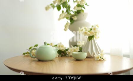 Traditionelles grünes Teeservice mit Keramik-Teekanne, kleinen Tassen und Vase mit weißen Blumen auf Holztablett, friedliche Stillleben-Komposition. Stockfoto