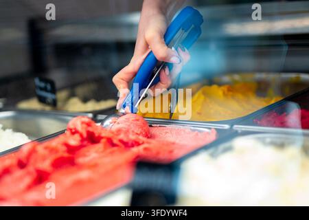 Hand schöpft lebendiges rotes Gelato mit blauem Metalllöffel aus gekühlter Vitrine. Stockfoto