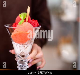 Hand präsentiert ein geschichtetes Dessert. Sahne, Pfirsich und rote Sorbet-Sphären füllen ein hohes Glas. Stockfoto