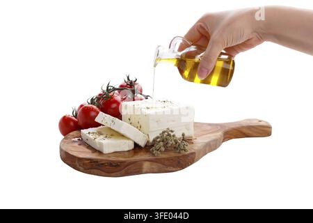 Weibliche Hand gießt Olivenöl auf griechischen Fetakäse mit Oregano und Tomaten, isoliert auf weißem Hintergrund Stockfoto