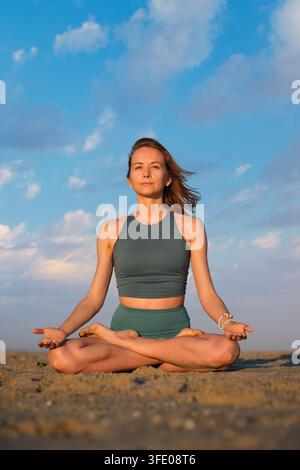 Mit offenen Augen und nach oben gerichteten Handflächen kniend sitzt eine Frau in Padmasana-Yoga, der Lotushaltung, und trägt grüne Sportkleidung an einem sandigen Strand Stockfoto