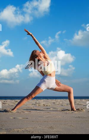 In einer halbgebundenen Viparita Virabhadrasana-Yoga-Variante, Reverse Warrior, streckt eine Frau einen Arm über den Kopf und legt den anderen hinter ihren Rücken am Strand Stockfoto