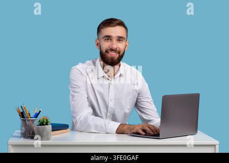 Millennial Geschäftsmann mit Laptop an seinem Schreibtisch, online arbeiten im Büro, blauen Studio Hintergrund Stockfoto
