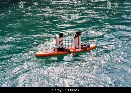 Zwei Personen paddeln gemeinsam auf einem leuchtend orangefarbenen Brett. Das Sonnenlicht tanzt über das türkisfarbene Wasser darunter. Sie lächeln und genießen ruhiges Wasser und klaren Himmel Stockfoto