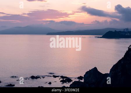 Weicher pastellfarbener Sommerabend über ruhigem Meer mit fernen dunklen Hügeln, die eine friedliche Küstenstimmung erzeugen Stockfoto