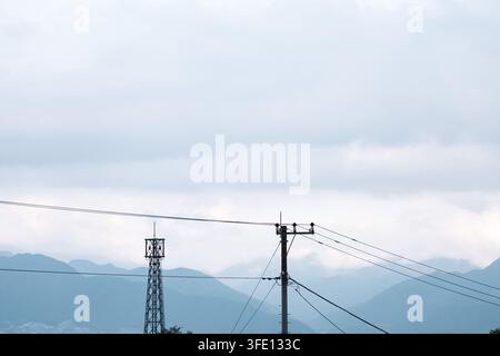 Ein Strommast und ein Gittermast für die Stromübertragung stehen als Silhouetten vor einem bewölkten Himmel mit geschichteten Bergen am Horizont in Japan und lassen oben einen weitläufigen negativen Raum. Stockfoto