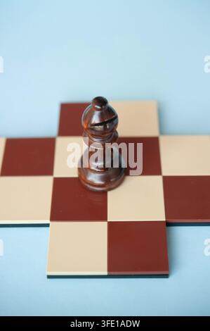 Nahaufnahme eines dunklen hölzernen Bischofs, der auf einem kleinen rot-cremefarbenen Schachbrett steht, vor einem hellblauen Hintergrund Stockfoto