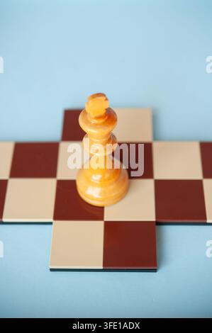 Nahaufnahme eines hellen hölzernen Schachkönigs, der auf einem kleinen roten und cremefarbenen Schachbrettkreuz vor einem hellblauen Hintergrund steht Stockfoto