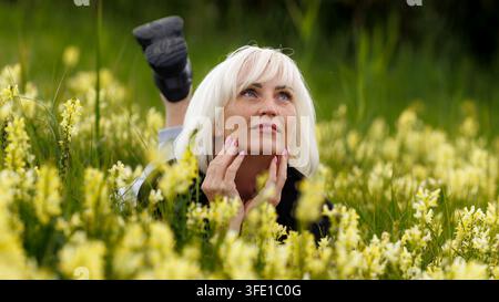 Ein Lifestyle-Porträt einer ruhigen blonden Frau, die in einer grünen Wiese voller gelber Wildblumen liegt, ihr Gesicht sanft berührt und nach oben schaut tho Stockfoto