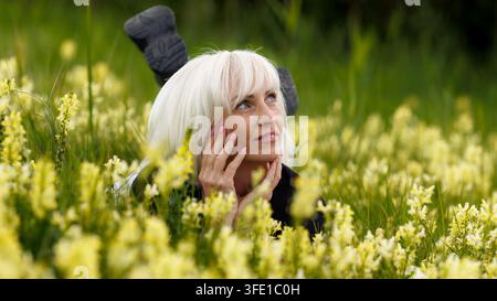 Ein Nahaufnahmeporträt einer nachdenklichen blonden Frau, die in einer üppigen Wiese liegt, umgeben von blühenden gelben Wildblumen, und einen friedlichen Moment genießt Stockfoto