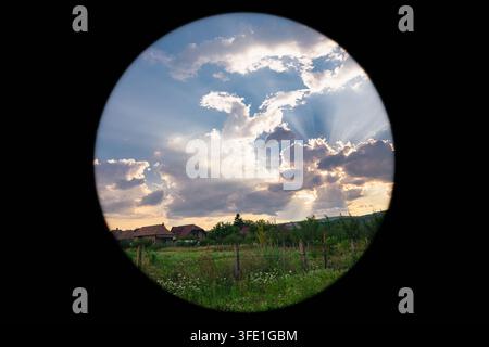 Sonnenuntergangshimmel über einem ländlichen Dorf, gesehen durch ein kreisförmiges Fenster mit dramatischen Wolken und Sonnenstrahlen im zentralen Transsilvanien, Rumänien. Stockfoto