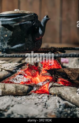 Traditioneller schwarzer Kessel, der über glühenden Kohlen und brennendem Feuerholz in einem rustikalen Kochbereich erhitzt wird Stockfoto