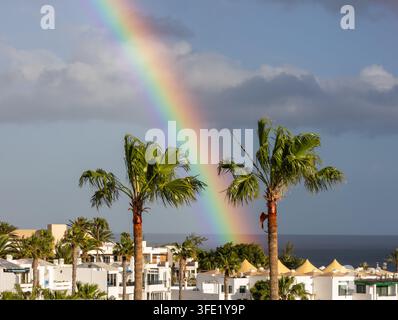 Detaillierte Ansicht eines lebhaften Regenbogens, der hinter Palmen und weißen Resortgebäuden an der Küste von Costa Teguise, Lanzarote, Spanien, aufsetzt Stockfoto