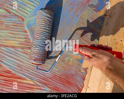 Hand führt Walze über lebendige, strukturierte Oberfläche. Farben vermischen sich dynamisch und erzeugen kräftige abstrakte Muster. Sonnenlicht hebt Kanten und Streifen hervor Stockfoto