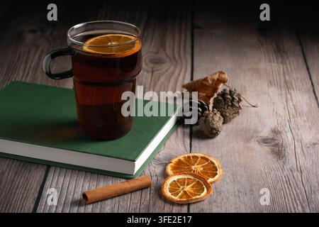 Wärmender, kuscheliger Tee mit einer Orangenscheibe, die auf einem grünen Buch ruht. Rustikale Holzoberfläche verziert mit Tannenzapfen, Herbstblättern und Gewürzen Stockfoto