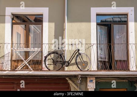 Schwarzes Vintage-Fahrrad, das auf einem schmalen Balkon zwischen zwei Glastüren mit Vorhängen an einer hellgrünen Wohnhausfassade in León, Spanien, geparkt ist. Warmes Sonnenlicht Stockfoto