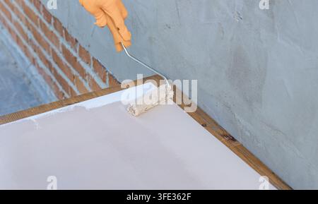 Hand trägt frische weiße Farbe auf einen Holzrahmen auf. Die Wand geht von grauer zu sauberer weißer Oberfläche über Stockfoto