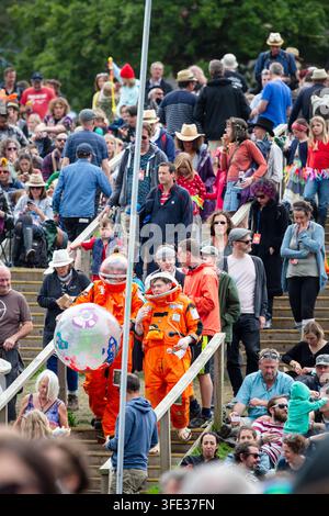 ASTRONAUTEN IN DER MENGE AUF DEM GREEN MAN FESTIVAL, REPORTAGE, GREEN MAN FESTIVAL 2017, BRECON, WALES, UK, 2017: Zwei Personen in leuchtend orangefarbenen Astronautenkostümen, eine hält einen aufblasbaren Globus, gehen über Holzstufen durch das sitzende Publikum auf einem Open-Air-Festival. Foto: Rob Watkins. INFO: Das Green Man Festival begann 2003 in Wales. Es findet jährlich an dem dritten Wochenende im August in den Brecon Beacons statt und feiert alternative Musik, Kunst, Literatur, Wissenschaft und familienfreundliche Kultur in einer wunderschönen Berglandschaft. Stockfoto