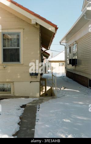 Eine schmale schneebedeckte Gasse zwischen zwei alternden holzverschalten Häusern an einem sonnigen Wintertag, mit einem geräumten Gehweg und einem sichtbaren Kellereingang. Stockfoto