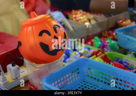 Orangen Plastik-Kürbislaterne-Eimer neben Körben mit bunten Spielzeugen und Packungen mit Süßigkeiten auf einem Nachtmarktstand. Stockfoto
