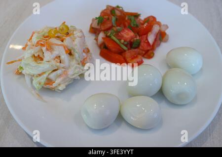 Ein weißer Essteller mit cremigem Krautsalat, gewürfelter Tomatensalsa und mehreren geschälten Wachteleiern, die in separaten Portionen angeordnet sind. Stockfoto