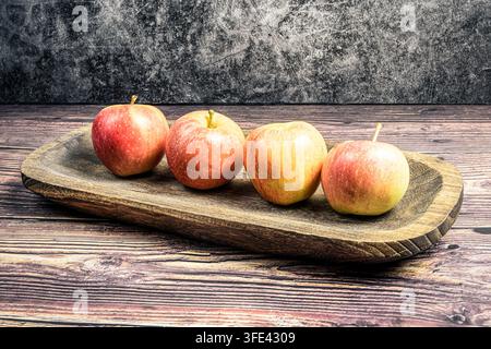 Vier rote und gelbe Gala-Äpfel, die auf einem länglichen rustikalen Holzteller aufgereiht sind, der auf einem Holztisch platziert wurde, vor einem grauen strukturierten Hintergrund. Stockfoto