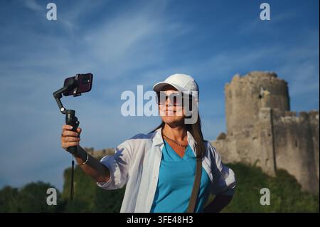 Lächelnde Reisende nimmt sich selbst mit einem Smartphone-Gimbal im Freien auf, mit einer alten Steinfortifikation und blauem Himmel im Hintergrund. Wales, Vereinigtes Königreich Stockfoto