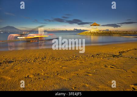 Traditionelles balinesisches Jukung-Außenbordboot am Strand bei Sonnenuntergang vertäut, mit einem steinernen Landungssteg-Pavillon und dem Vulkan Mount Agung unter einem sternenklaren Nachthimmel, Sanur, Bali. Stockfoto