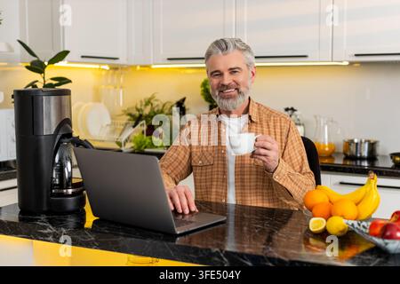 Ein Mann mittleren Alters arbeitet an einem Laptop in der Küche und trinkt heißen Kaffee, während er E-Mails für freiberufliche Fernarbeit liest. Reifer Kerl fühlt sich glücklich produktiv während der Pause und hält weiße Tasse in der modernen Küche. Stockfoto