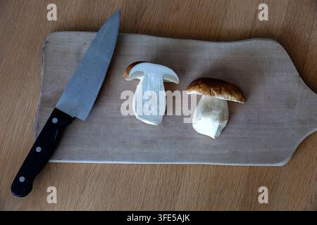 Eine detaillierte Nahaufnahme eines frischen Porchininpilzes, der auf einem rustikalen Holzschneidebrett halbiert wurde, mit einem professionellen Küchenmesser daneben. Stockfoto