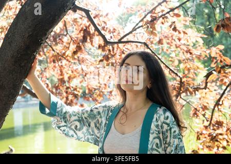 Porträt einer attraktiven Frau, die einen Baumstamm im Park in der Nähe eines Teiches berührt Stockfoto