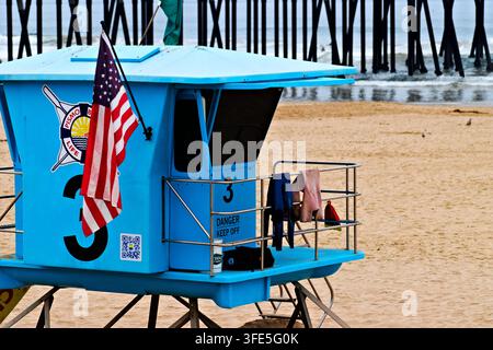 Heller blauer Rettungsturm mit einer amerikanischen Flagge steht auf einem Sandstrand neben Pierpfählen und Ozeanwellen in Pismo Beach, Kalifornien. Stockfoto