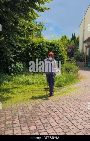 Ein Mann in einem grauen Hemd und orangefarbener Sicherheitsausrüstung geht auf einem Steinweg. Er verwendet ein Rasenmäherwerkzeug in einem Garten mit grünen Pflanzen drumherum. Die Sonne scheint Stockfoto