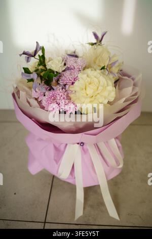 Pastellfarbenes Blumenbouquet mit weißen Nelken, violetten Chrysanthemen und Clematis, elegant in Lavendelpapier mit weißen Bändern verpackt für Hochzeiten und Feiern Stockfoto