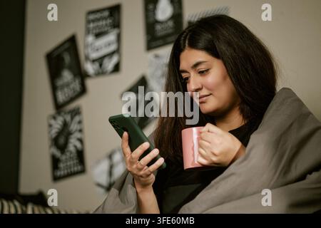Bikolor Frau hält Tasse und benutzt Smartphone im Bett Stockfoto