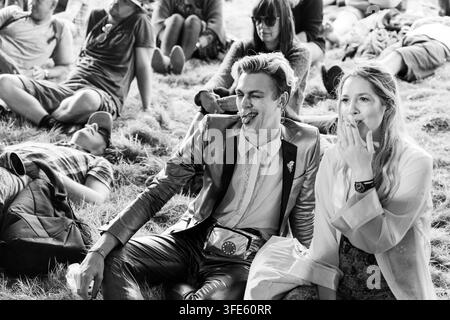 FRECHES PAAR IN GLAM-OUTFITS AUF EINEM FESTIVAL IN SCHWARZ-WEISS, REPORTAGE, GREEN MAN FESTIVAL 2018, BRECON, WALES, UK, 2018: Ein stilvoller junger Mann in einer glänzenden Jacke zwinkert und streckt die Zunge heraus, während er im Gras neben einer rauchenden Frau sitzt, in diesem Schwarz-Weiß-Schnappschuss. Foto: Rob Watkins. INFO: Das Green Man Festival begann 2003 in Wales. Es findet jährlich an dem dritten Wochenende im August in den Brecon Beacons statt und feiert alternative Musik, Kunst, Literatur, Wissenschaft und familienfreundliche Kultur in einer wunderschönen Berglandschaft. Stockfoto