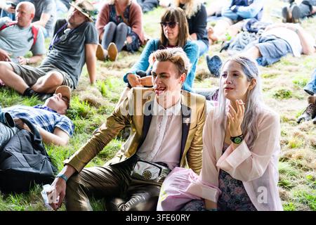 STILVOLLES PAAR IN GLAM-OUTFITS ENTSPANNT AUF EINEM FESTIVAL, REPORTAGE, GREEN MAN FESTIVAL 2018, BRECON, WALES, UK, 2018: Ein stilvoller junger Mann in einer goldenen Jacke und glänzenden Hosen zwinkert und streckt die Zunge heraus, während er auf dem Gras neben einer rauchenden Frau sitzt. Foto: Rob Watkins. INFO: Das Green Man Festival begann 2003 in Wales. Es findet jährlich an dem dritten Wochenende im August in den Brecon Beacons statt und feiert alternative Musik, Kunst, Literatur, Wissenschaft und familienfreundliche Kultur in einer wunderschönen Berglandschaft. Stockfoto