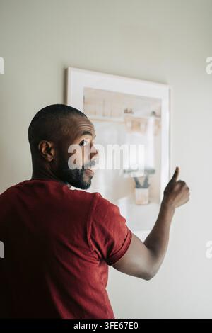 Mann schaut über die Schulter und macht Daumen-hoch-Geste, während er ein Gemälde an seiner neuen Wohnung an die Wand hängt Stockfoto