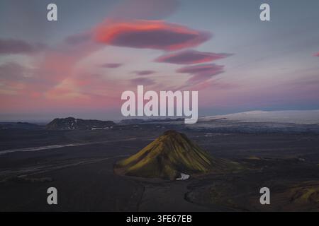 Luftaufnahme des Vulkans Maelifell mit lebendigen grünen, moosbedeckten Hängen, weite vulkanische Landschaft, entfernter Gletscher und dramatische rosa Linsenwolken Stockfoto