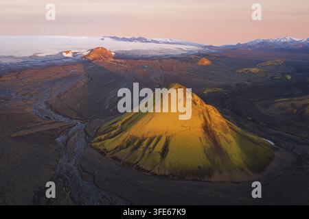 Luftaufnahme des Maelifell-Vulkans mit seinen lebendigen moosbedeckten Hängen vor einer weiten vulkanischen Landschaft und einem fernen Gletscher bei weicher Goldener Stunde Stockfoto
