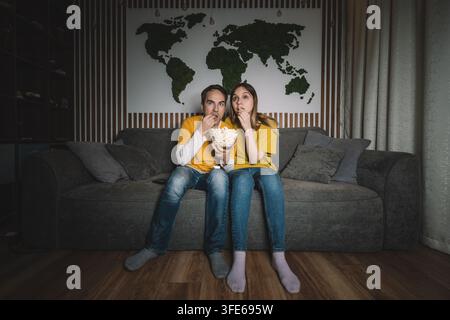 Paar sitzt abends zu Hause auf einem Sofa und schaut einen interessanten Film mit Popcorn. Gemütlicher Familienabend, Kinoatmosphäre und gemeinsame Unterhaltung Stockfoto