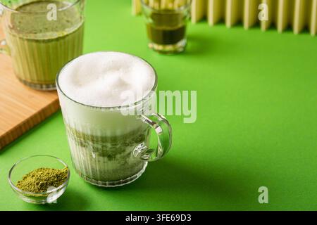 Matcha-Latte in einer klaren Glastasse mit schaumiger Milch, begleitet von einer kleinen Schüssel Matcha-Pulver auf einem lebendigen grünen Hintergrund Stockfoto